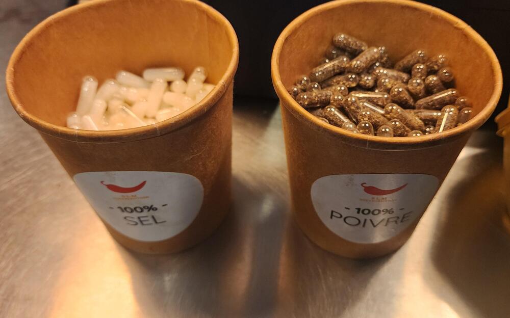 Two brown cups labeled “100% Sel” and “100% Poivre” contain white and dark brown capsules, respectively. The cups are placed side by side on a metallic surface.
