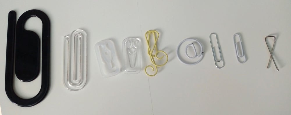 Paper clips of various shapes, sizes, and colors are arranged in a row on a white surface. Each clip has a unique design, including a musical note and an “@” symbol.