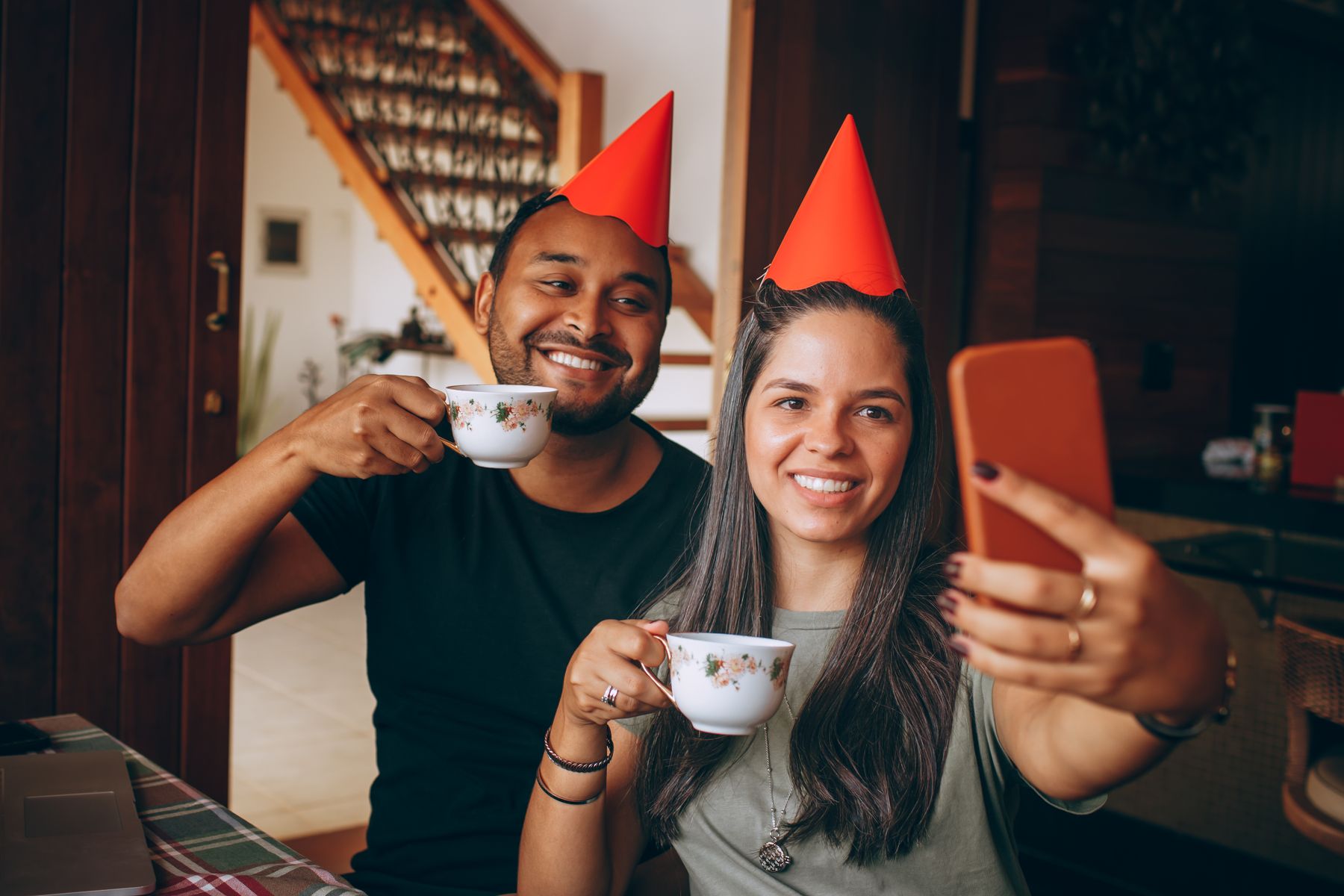 Two people wearing red party hats smile while holding teacups and taking a selfie together indoors. They are sitting at a table, celebrating in a cozy home setting.