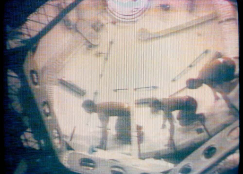 Two astronauts inside a spacecraft or space station module, working near the floor under dim lighting, with equipment and structural details visible around them. The image has a grainy, vintage TV appearance.