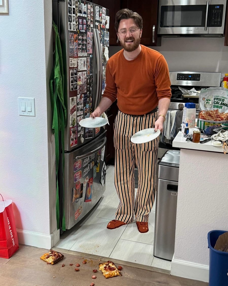 A man in striped pants and an orange shirt stands in a kitchen holding plates, looking surprised. Two slices of pizza have fallen face down on the floor, with toppings scattered around.