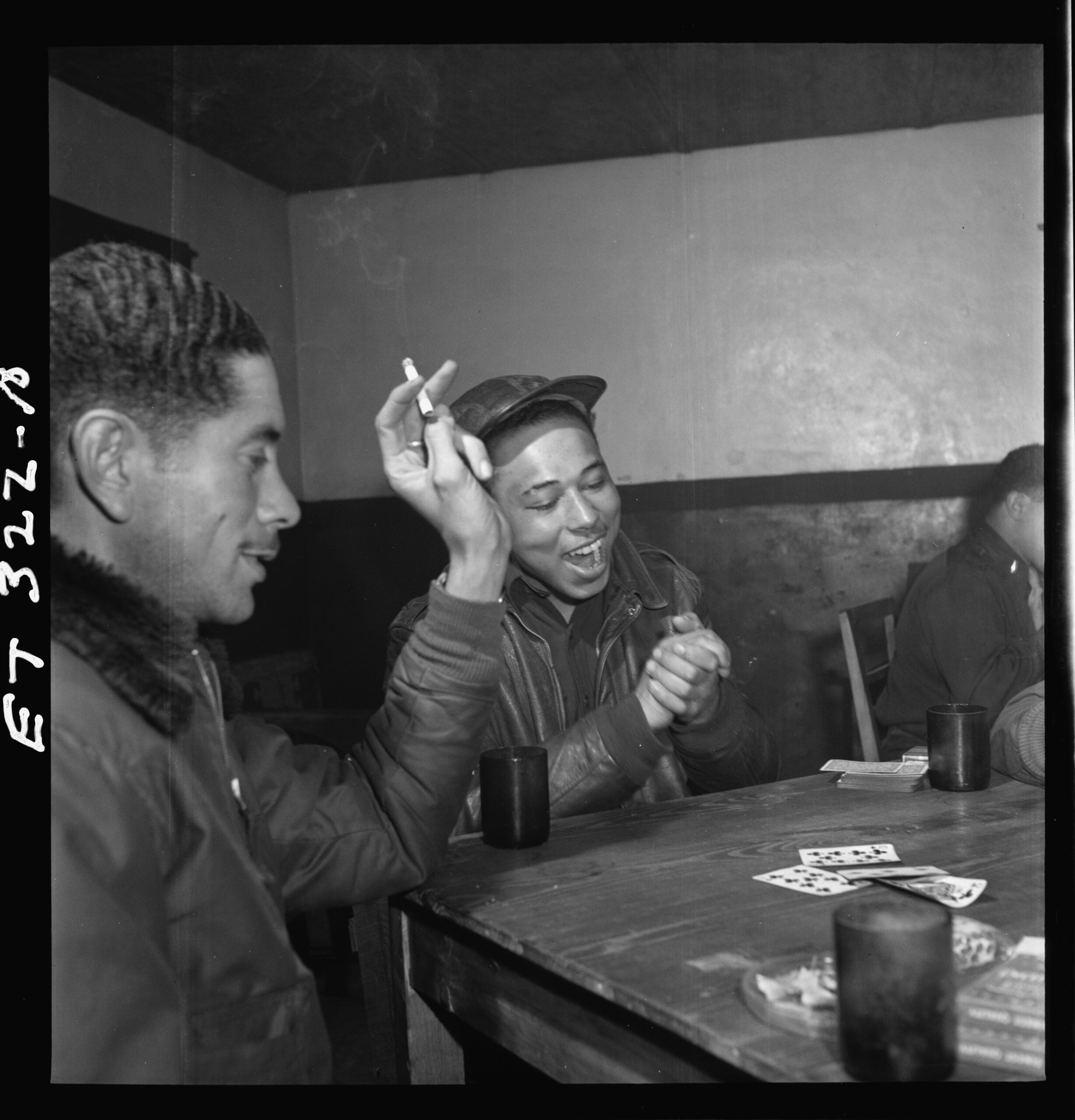 Three men sit at a table playing cards. One man is smiling and clapping, another holds a cigarette, and a third man is partially visible. Three cups sit on the wooden table. The mood appears relaxed and cheerful.
