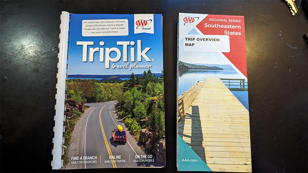 Two AAA travel guides are on a black surface: one has a cover showing a car on a scenic road, and the other is a folded map labeled “Southeastern States TRIP OVERVIEW MAP.”