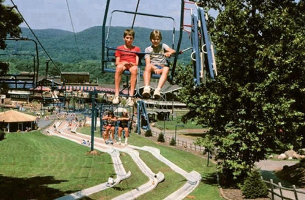 Two children ride a ski lift over a green hill with a winding alpine slide below. Other people are riding the slides, and trees and mountains are visible in the background on a sunny day.