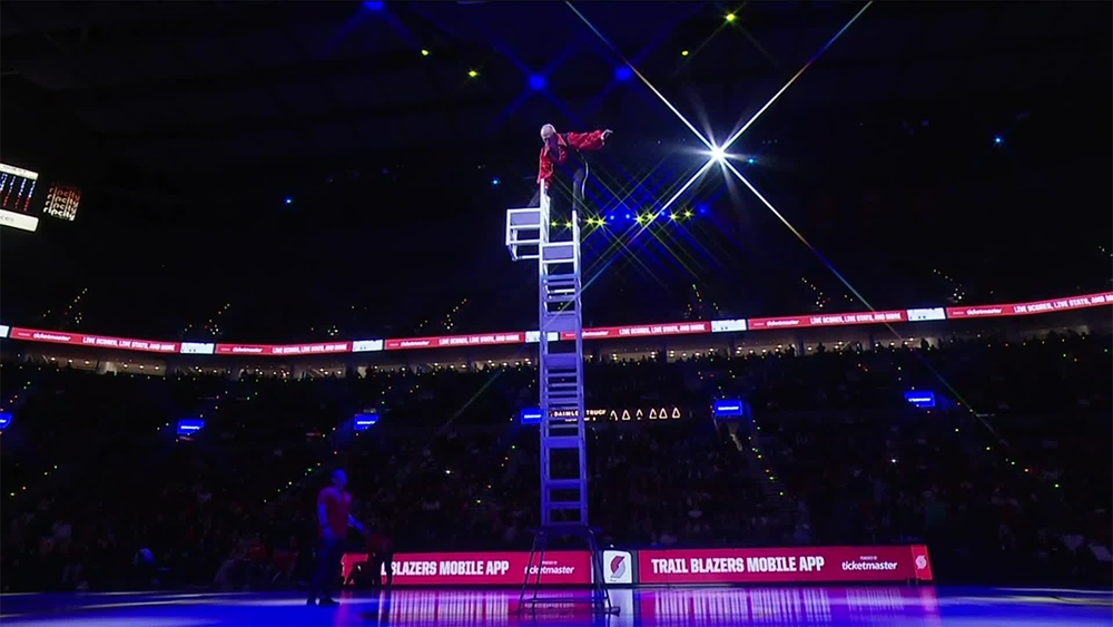 A performer in red balances atop a tall stack of chairs at center court in a darkened arena, with bright lights and a large audience watching.