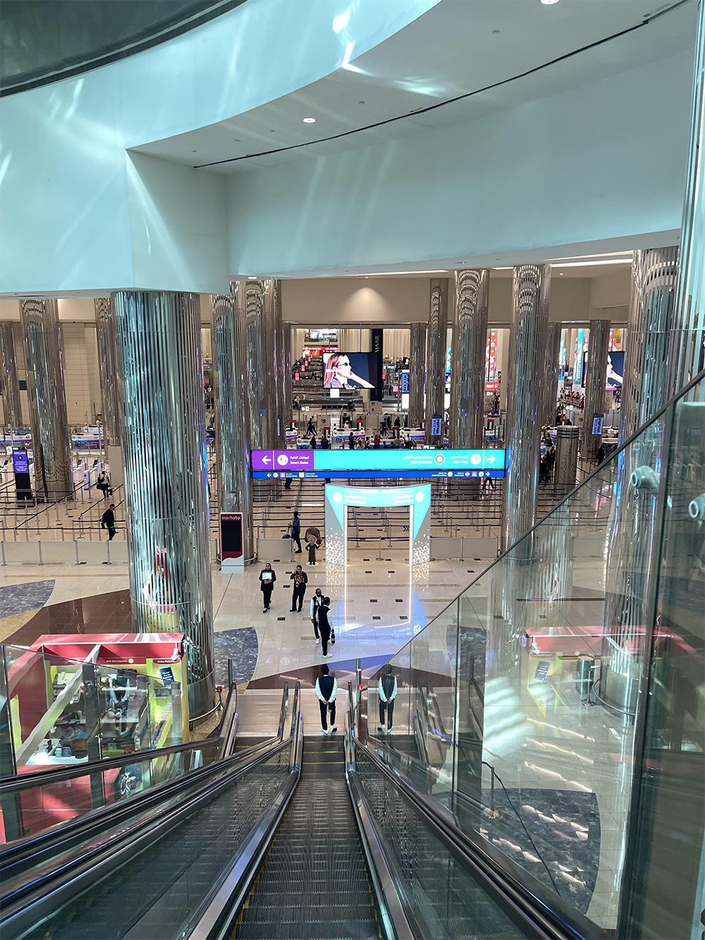View from an escalator inside a modern, brightly-lit airport terminal with shiny pillars, digital screens, and people walking near check-in areas.