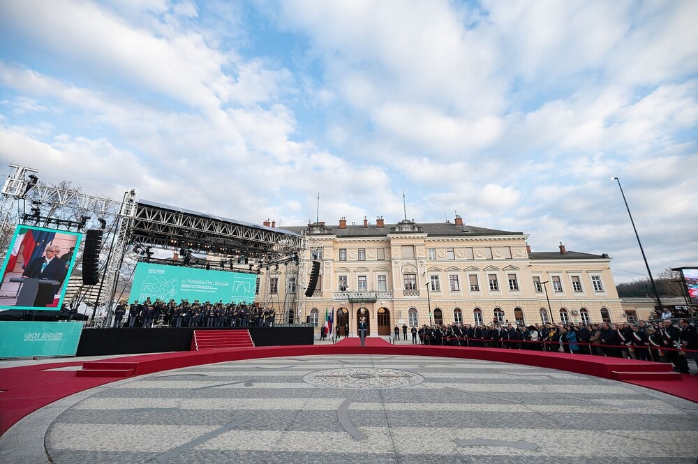 An outdoor ceremony with a large circular stage, red carpet, and audience in front of a grand historic building under a partly cloudy sky. A screen and musicians are visible on the left.