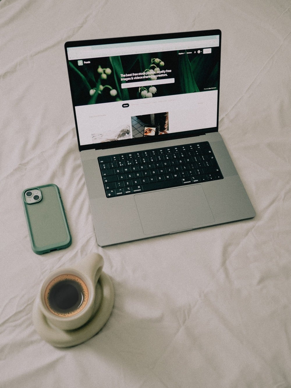 A laptop with a website open, a smartphone with a teal case, and a cup of coffee on a white bed sheet.