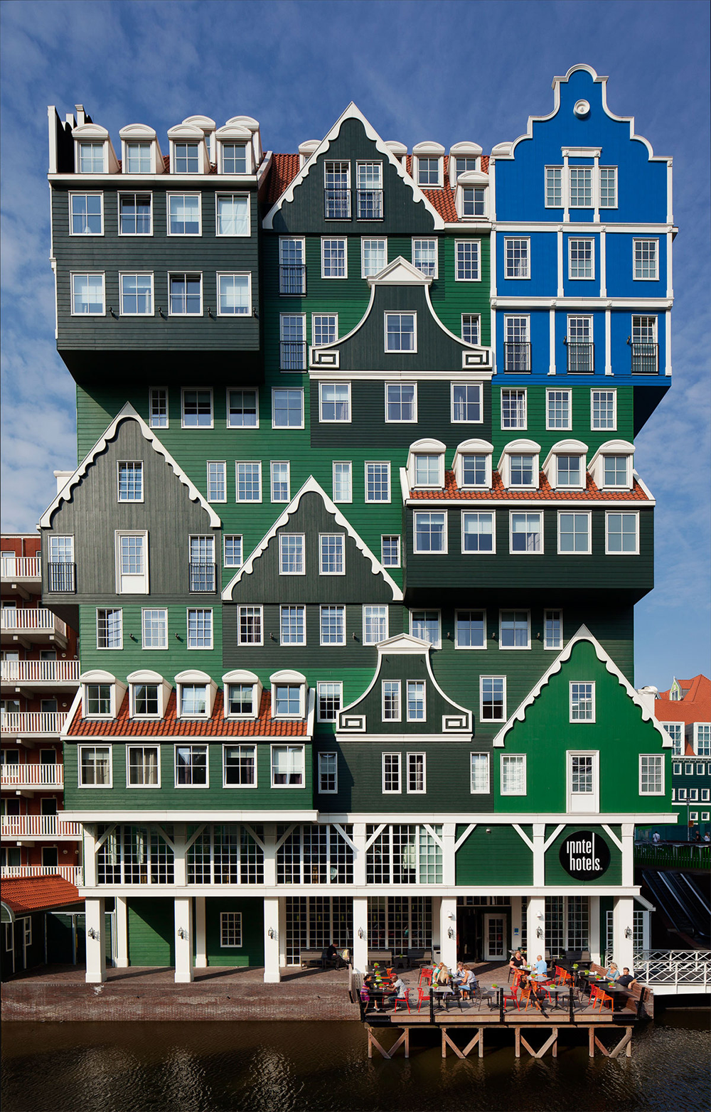 A quirky hotel with stacked green and blue house-like façades, white trim, and a cafe terrace by the water, under a sunny blue sky.