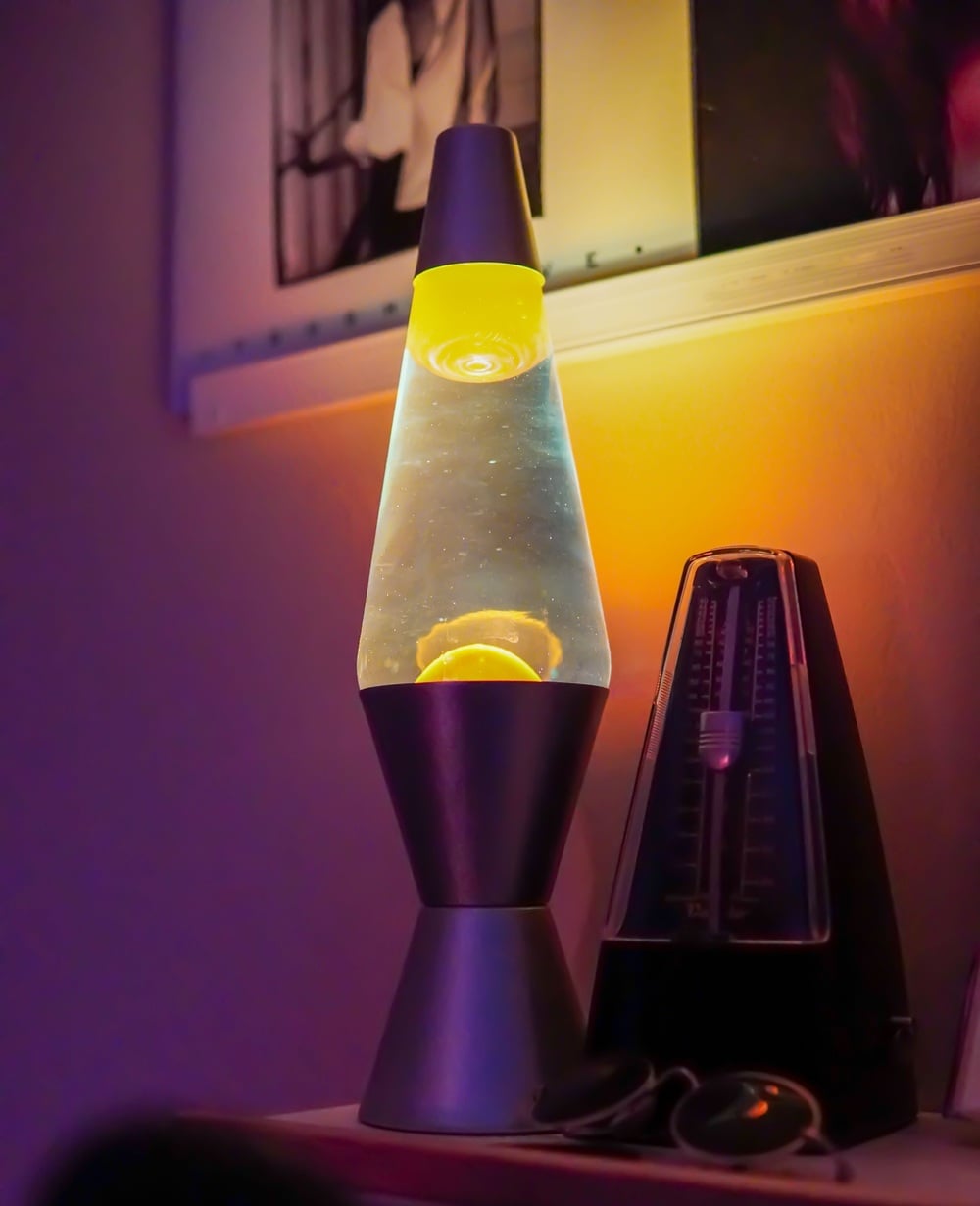 A glowing lava lamp with yellow wax sits on a shelf beside a black metronome, with blurred framed pictures hanging on the wall behind them. Warm, ambient lighting fills the scene.