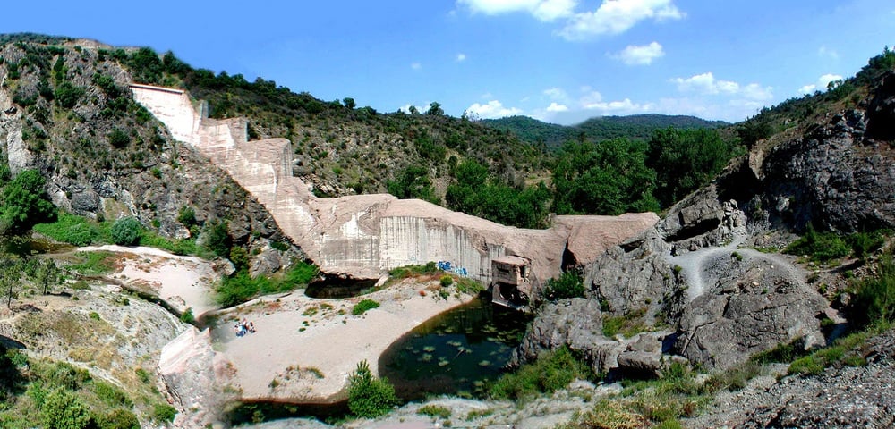 A stepped concrete dam is built into a rocky hillside with a small pool of water at its base, surrounded by greenery, rocks, and a sandy area where a few people are sitting. Blue sky with scattered clouds overhead.