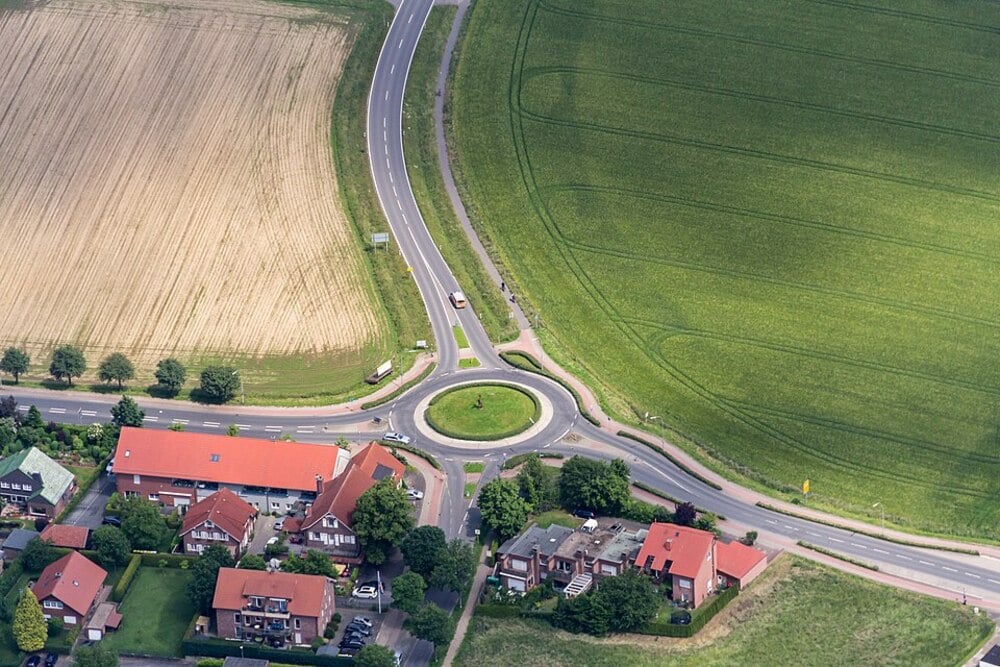 Aerial view of a roundabout surrounded by fields and houses, intersecting roads with vehicles, green grass on one side, plowed field on the other, and homes with red roofs nearby.