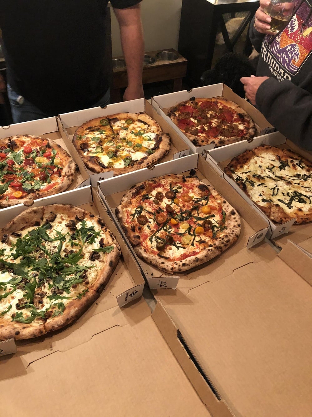 Six open pizza boxes with assorted gourmet pizzas on a table. Two people are standing nearby, one holding a drink. The pizzas have a variety of toppings including greens, vegetables, meats, and cheeses.