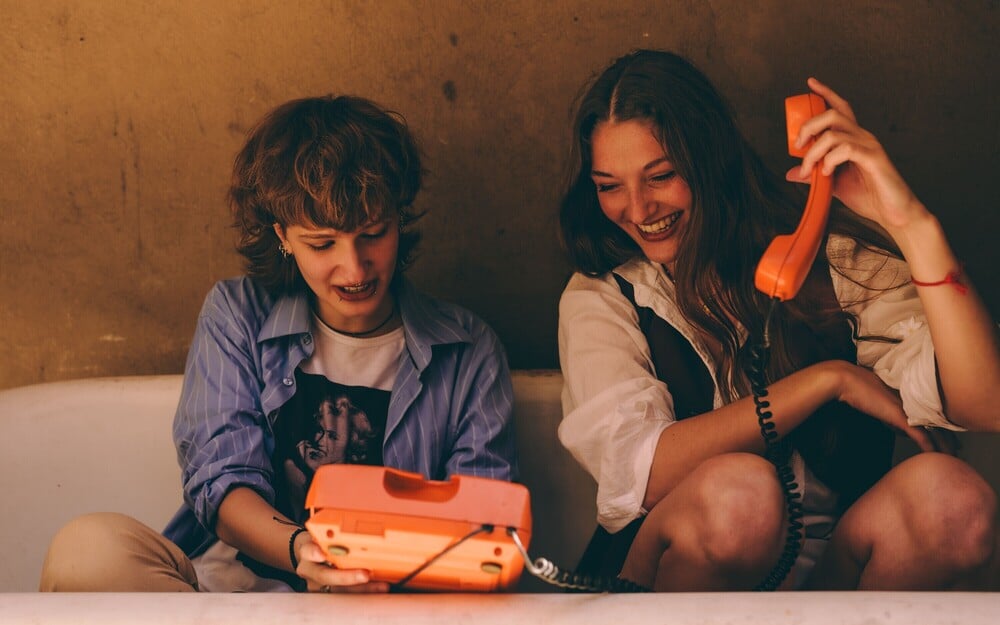 Two young people sitting in a bathtub, laughing while using an orange corded telephone. One holds the handset to their ear, while the other touches the phone base. Warm lighting creates a relaxed, playful atmosphere.