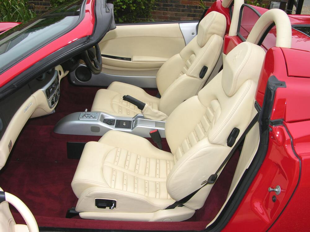 The image shows the interior of a red convertible sports car with cream leather seats, a center console, and a gear shifter. The car features a modern, luxurious design with clean lines and detailed seat stitching.