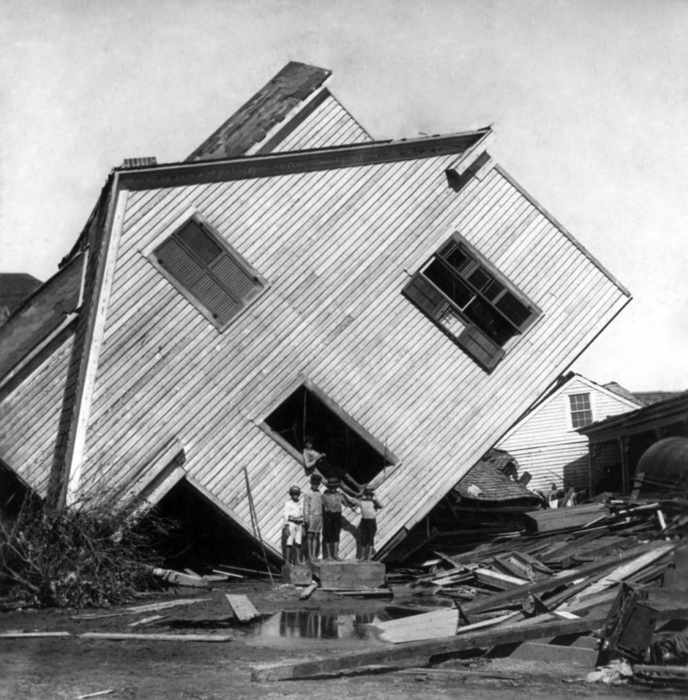 A group of children stand in front of a house tilted sideways, surrounded by debris and floodwater, after a natural disaster.