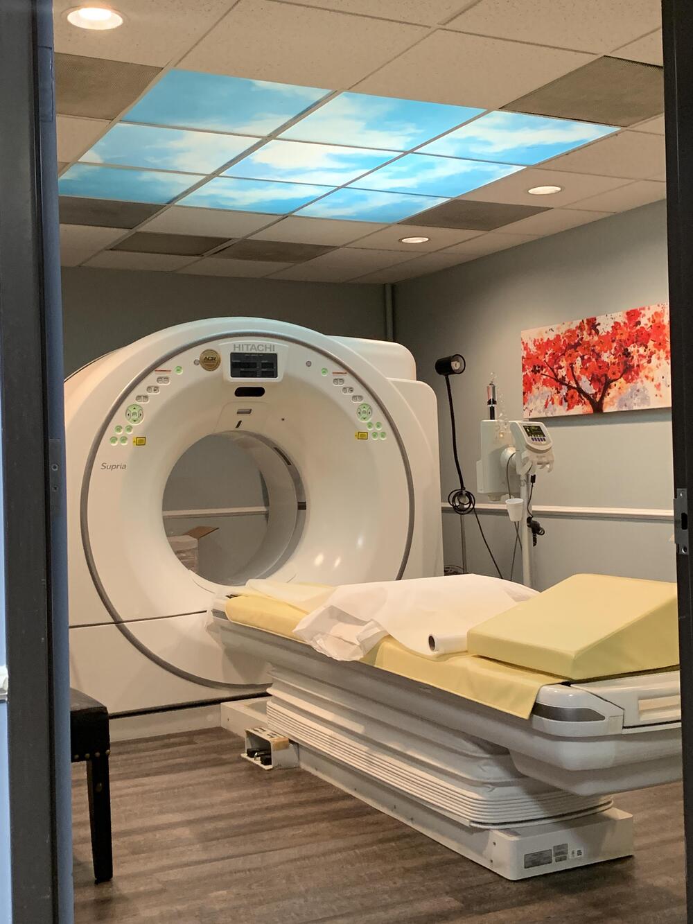 A CT scan machine with a yellow-cushioned table in a medical room. The ceiling has a light panel showing a sky scene, and a red tree artwork is on the wall.