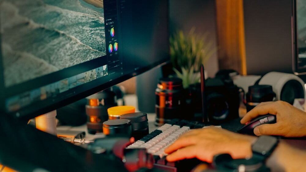 A person edits a photo of a beach on a computer with a large screen, surrounded by camera lenses and photography equipment on a cluttered desk.