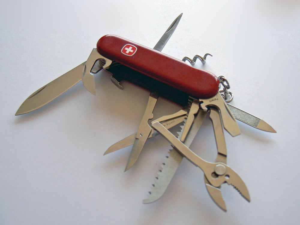 A red Swiss Army knife with multiple tools extended, including blades, saw, scissors, pliers, can opener, corkscrew, and screwdriver, on a white surface.