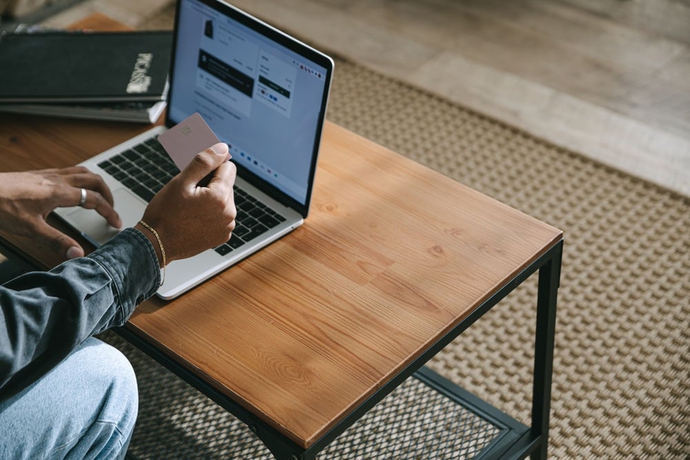 A person holds a credit card in one hand while using a laptop on a wooden table, possibly making an online transaction or purchase. A closed black notebook sits on the table next to the laptop.