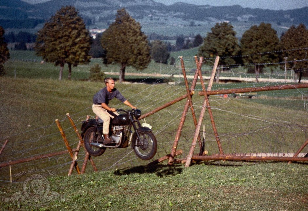 A man rides a motorcycle, jumping over a barbed wire fence in a grassy, rural landscape with trees and hills in the background.