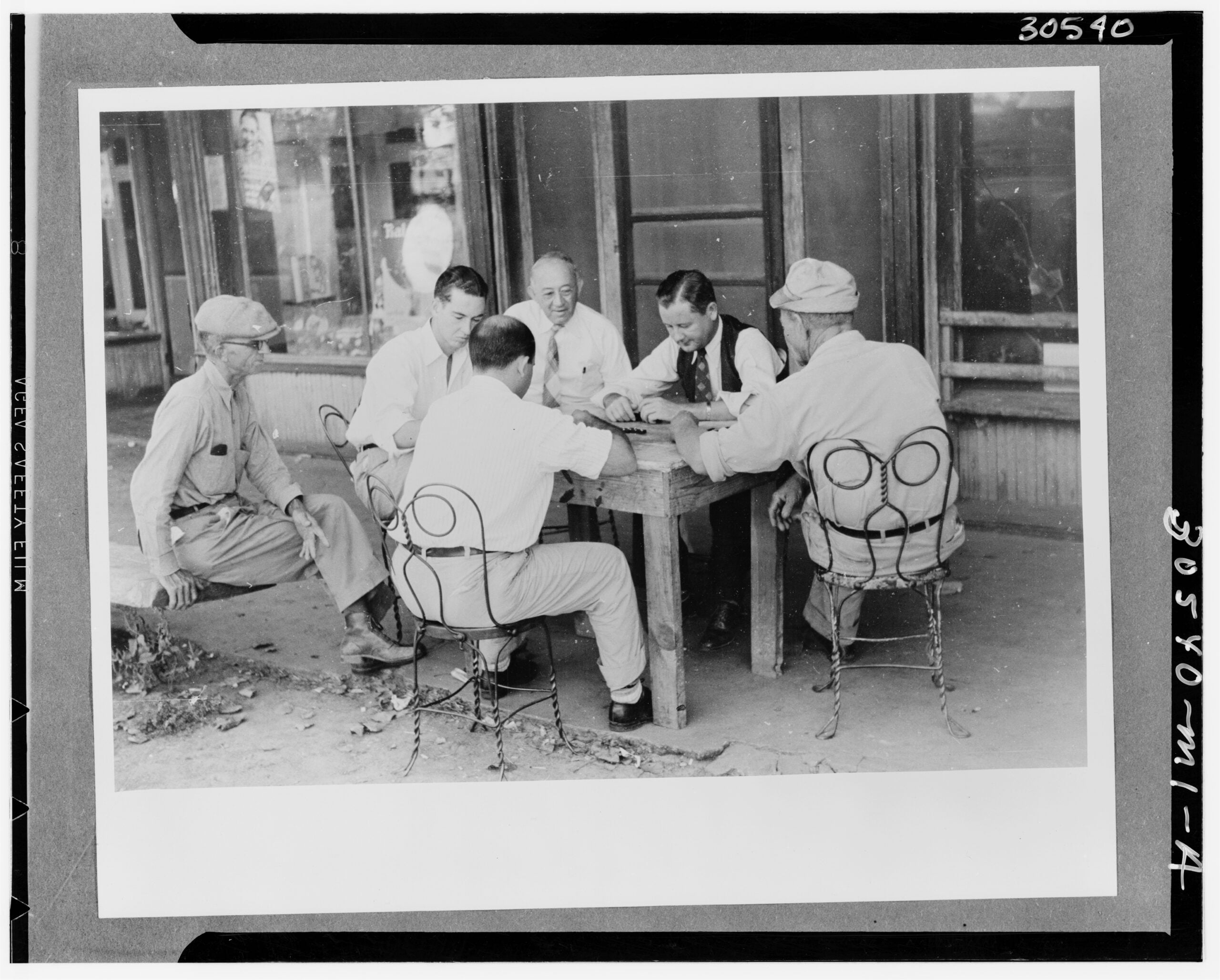 Five men sit around a wooden table outside, engaged in a card game. Two are in light-colored shirts and ties, three wear hats. They are seated on metal chairs on a sidewalk in front of a building with large windows.