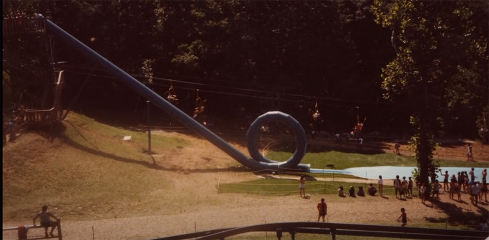 A blue waterslide with a full vertical loop ends in a small pool. People are waiting in line and watching, surrounded by grass and trees under sunlight.