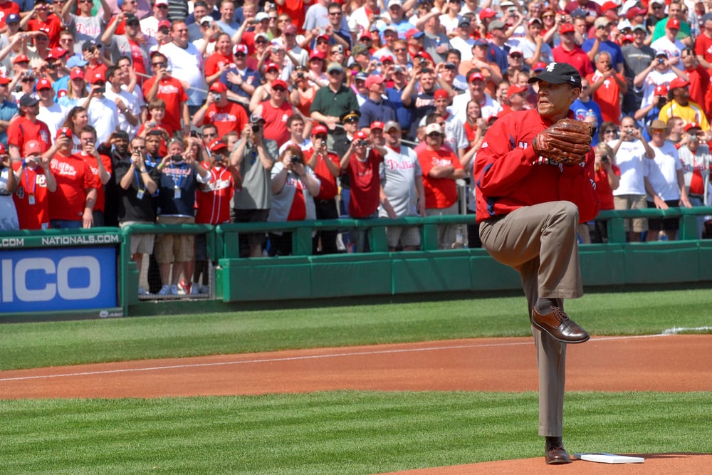 A man in a red jacket and baseball cap winds up to throw a ceremonial first pitch on a baseball field as a large crowd watches and takes photos in the stands behind him.