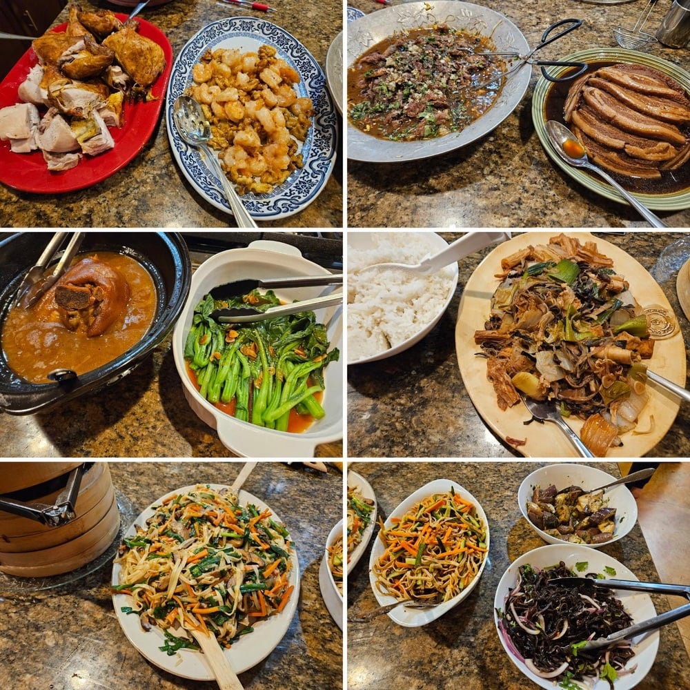 A variety of Chinese dishes, including meats, vegetables, rice, and noodles, are served on plates and bowls on a granite table, ready for a feast. Multiple serving utensils are placed with each dish.