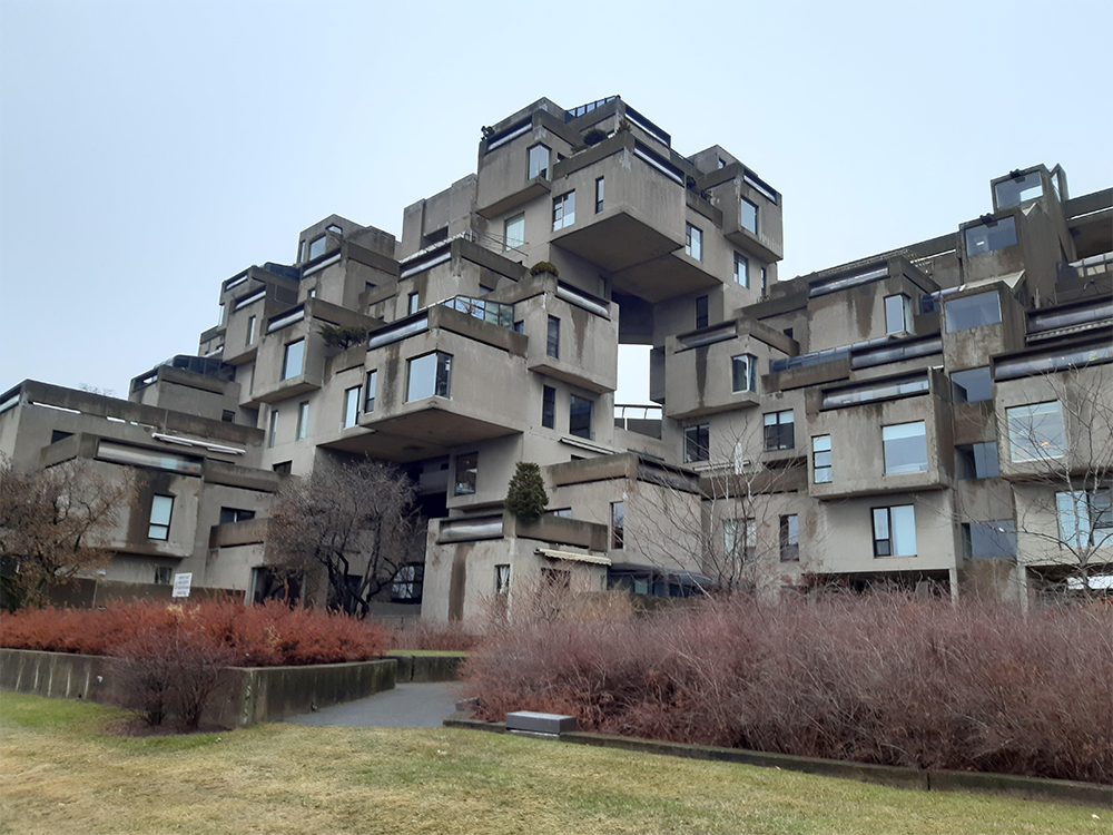 A unique building made of stacked, block-like concrete units with many windows, balconies, and terraces. Brown shrubbery and grass are in the foreground under a cloudy sky.