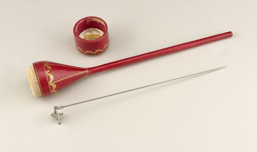 A red, gold-decorated sewing tool set including a thimble, a long needle with a charm, and a cylindrical case, all displayed on a white surface.