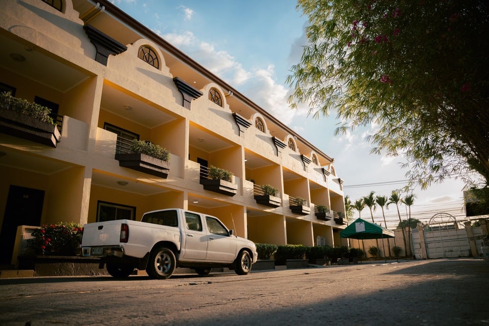 A white pickup truck is parked in front of a yellow, three-story building with balconies and potted plants, under a blue sky with scattered clouds.