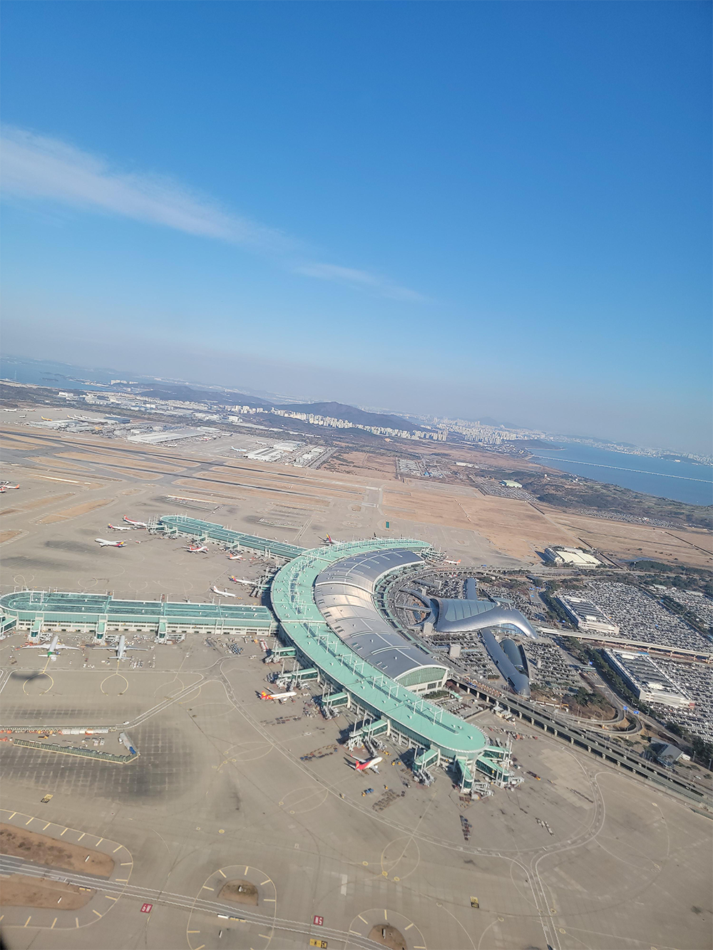 Aerial view of a large airport terminal with several airplanes parked, runways, and taxiways. The landscape includes parking lots, buildings, and distant city and coastline under a clear blue sky.