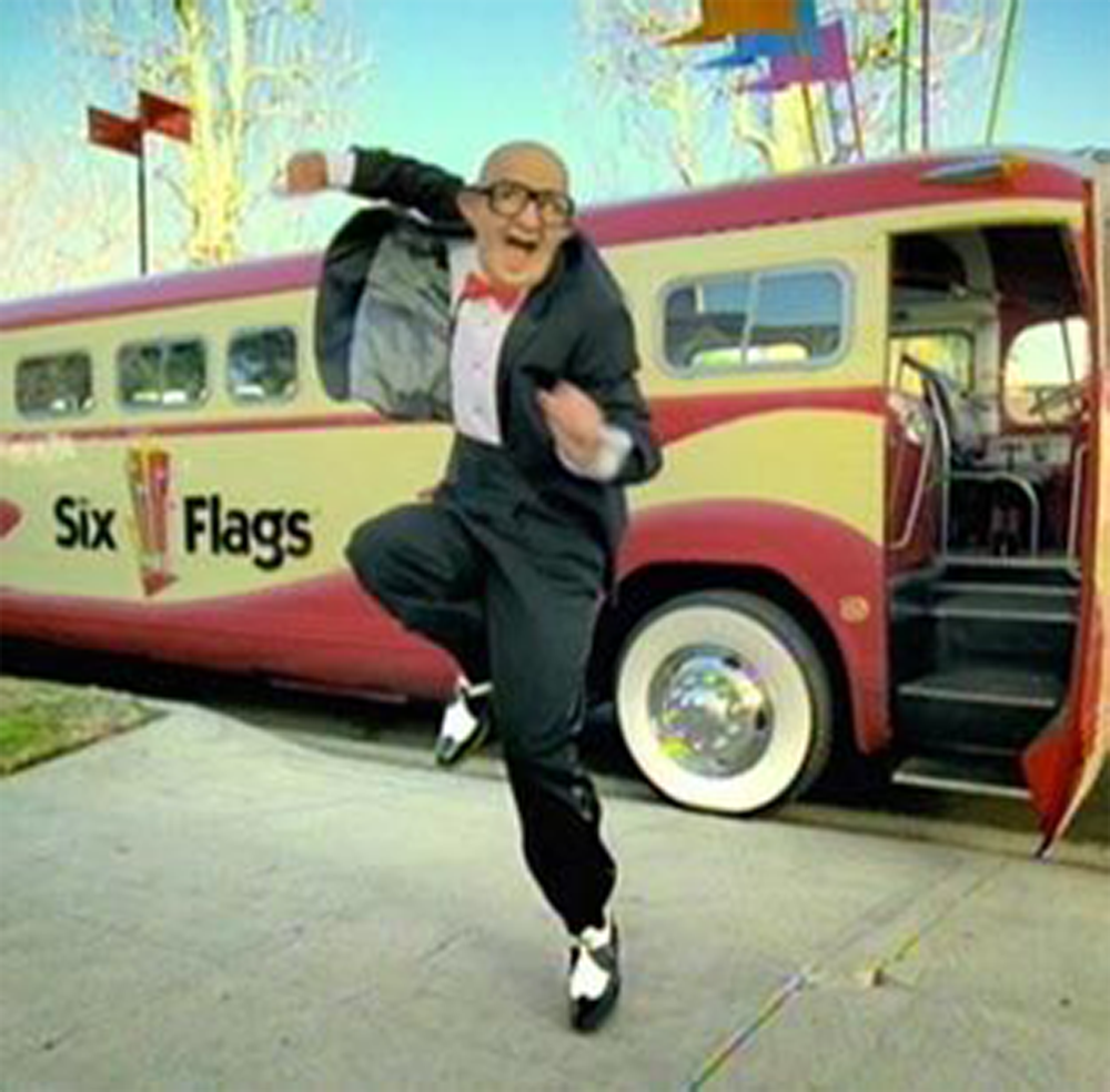 A man in a tuxedo and glasses energetically dances in front of a retro Six Flags bus parked on a sidewalk, with colorful flags in the background.