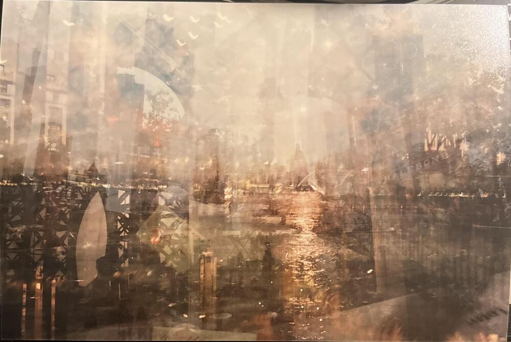 A hazy, artistic image showing a river at sunset with reflections on the water, city buildings, and bridge arches overlapping in a dreamy, layered composition.
