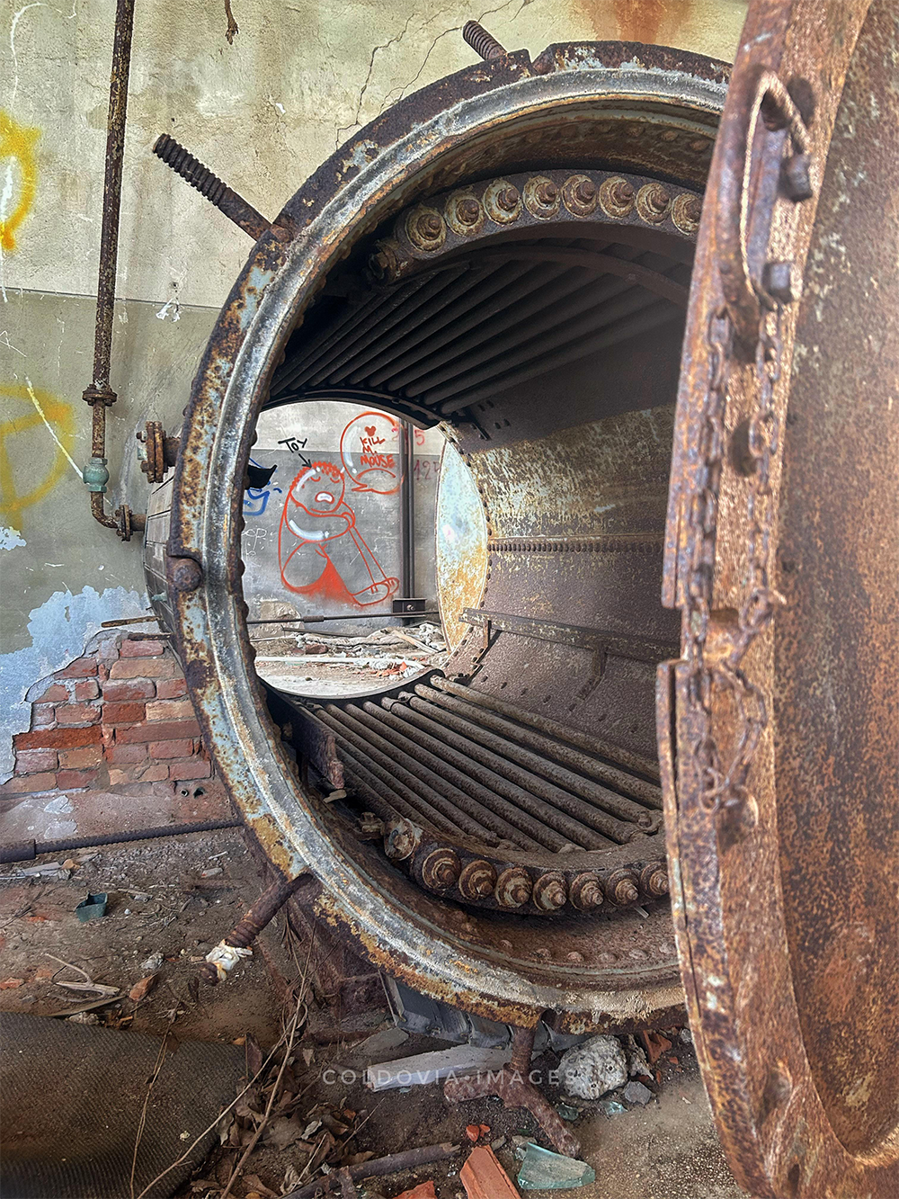 A large, rusty, cylindrical metal chamber with an open door sits in an abandoned building; graffiti and debris are visible through the opening.