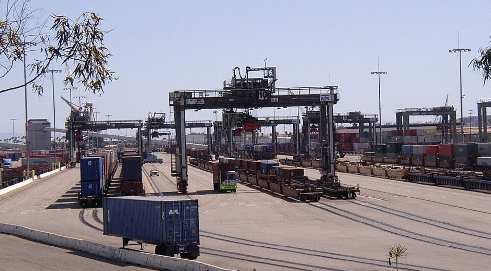 Shipping containers and cranes at a busy port terminal, with a truck and equipment moving cargo under clear skies.