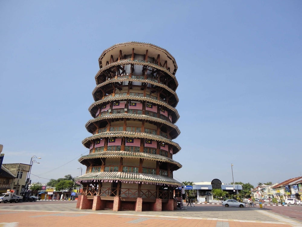 A multi-story leaning tower with Malaysian flags, located in an open square, surrounded by low-rise buildings under a clear blue sky.
