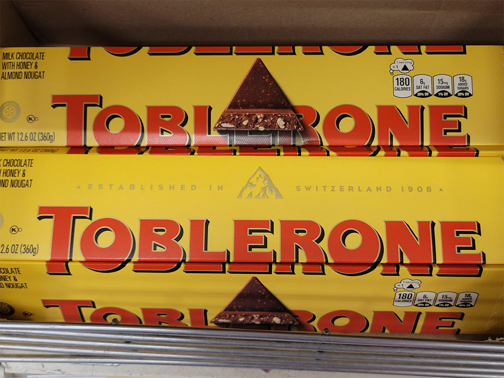 Two yellow Toblerone chocolate bars with red lettering are stacked on a shelf. The packaging shows a mountain graphic and nutritional information on the right side.