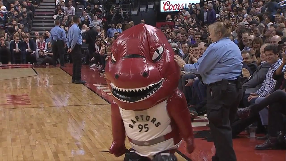 A person in a red dinosaur mascot costume wearing a Raptors jersey walks on a basketball court, with security and audience members in the background.