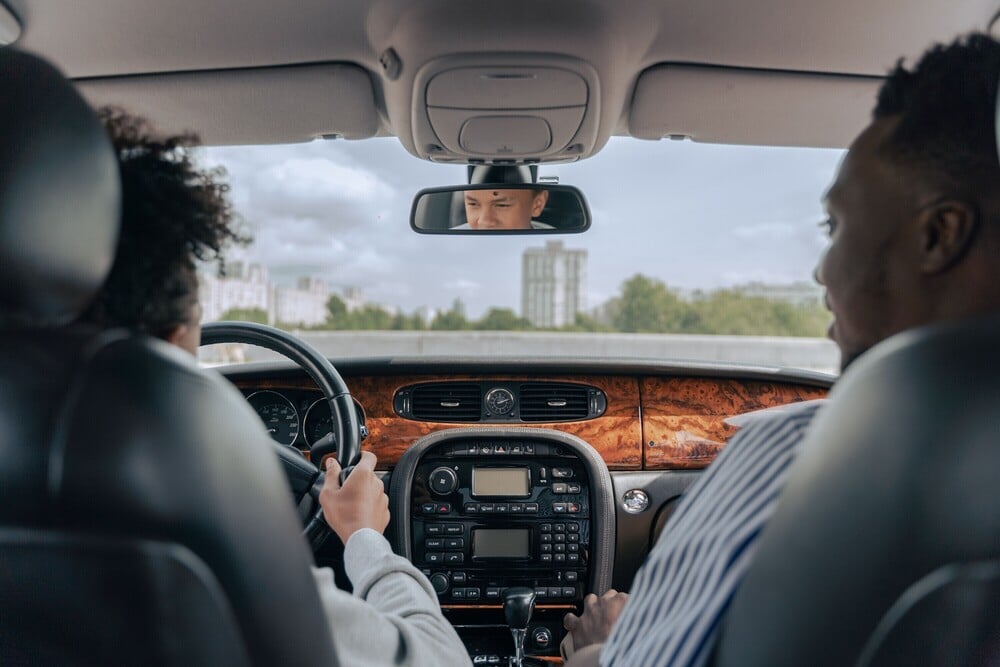 View from the backseat of a car with two people in the front seats and a person visible in the rearview mirror, driving through a city with buildings and trees seen through the windshield.