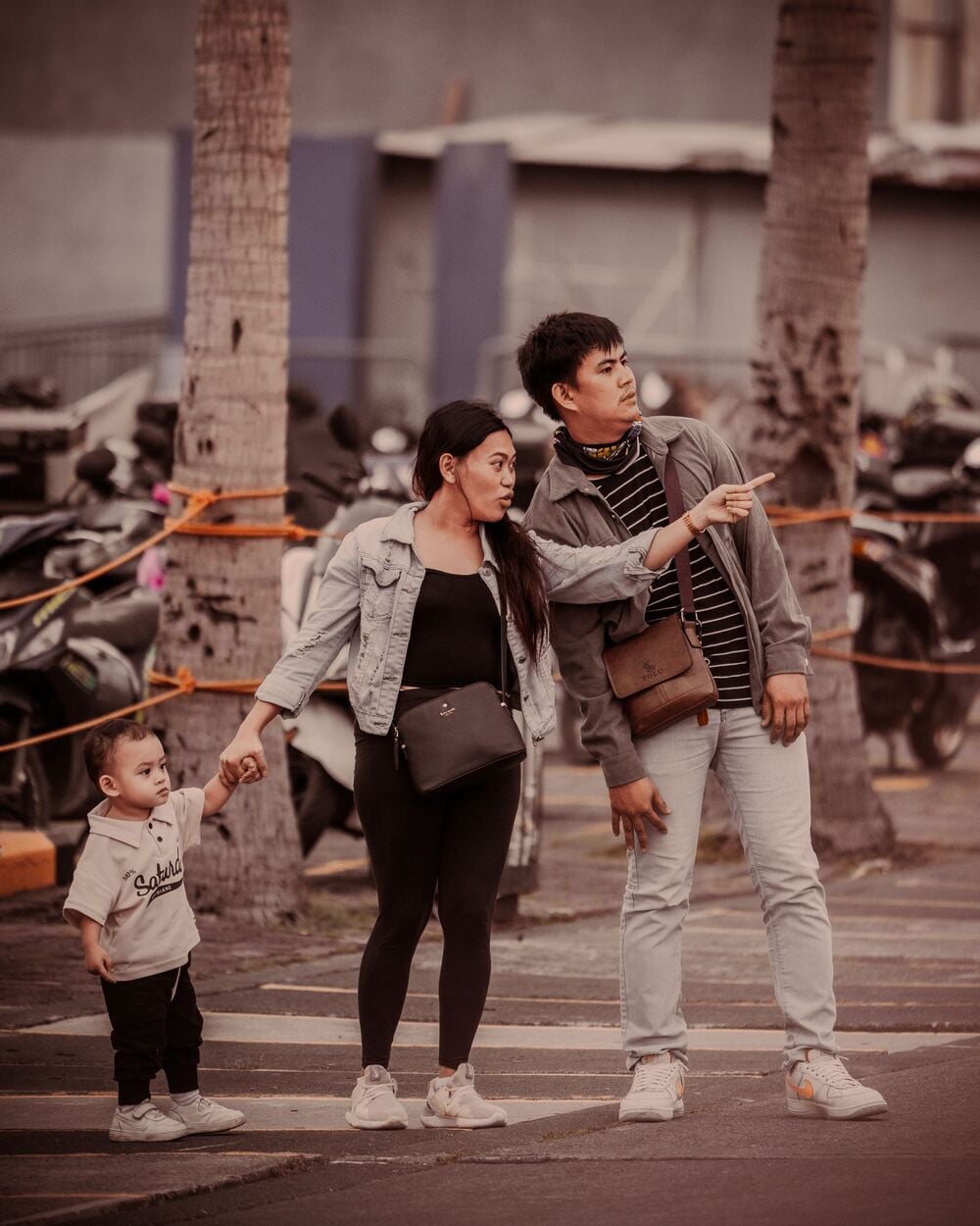 A family of three stands outdoors near palm trees and motorcycles. The parents, holding their child’s hand, both point in the same direction while the child looks elsewhere. All are dressed casually.