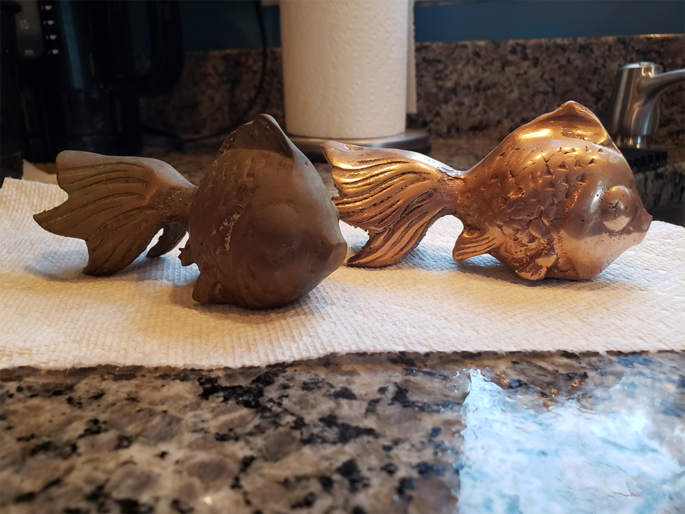 Two fish-shaped metal objects sit on a paper towel atop a granite counter. One fish is tarnished and dull, while the other is shiny and polished. A sink and other kitchen items are in the background.