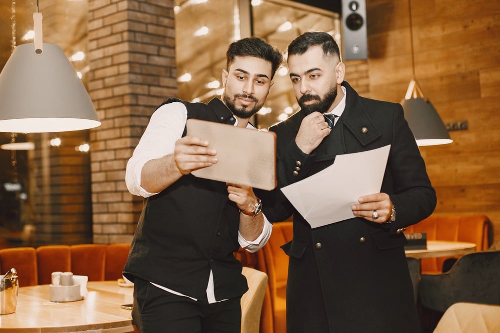 Two men in black suits stand in a cozy café, looking at a tablet and holding papers. One man is showing the screen to the other, who touches his chin thoughtfully. Warm lighting and wooden décor are in the background.