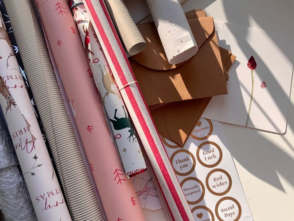 Several rolls of decorative wrapping paper, brown envelopes, a sheet with circular stickers, and a greeting card with a single red flower are arranged on a sunlit white surface.