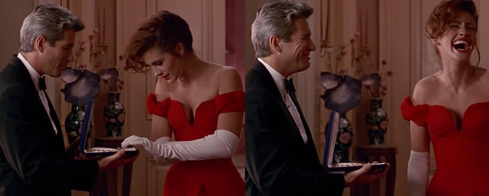 A man in a tuxedo surprises a woman in a red dress by snapping a jewelry box shut as she reaches inside, making her laugh. They both appear joyful and amused in an elegant room.