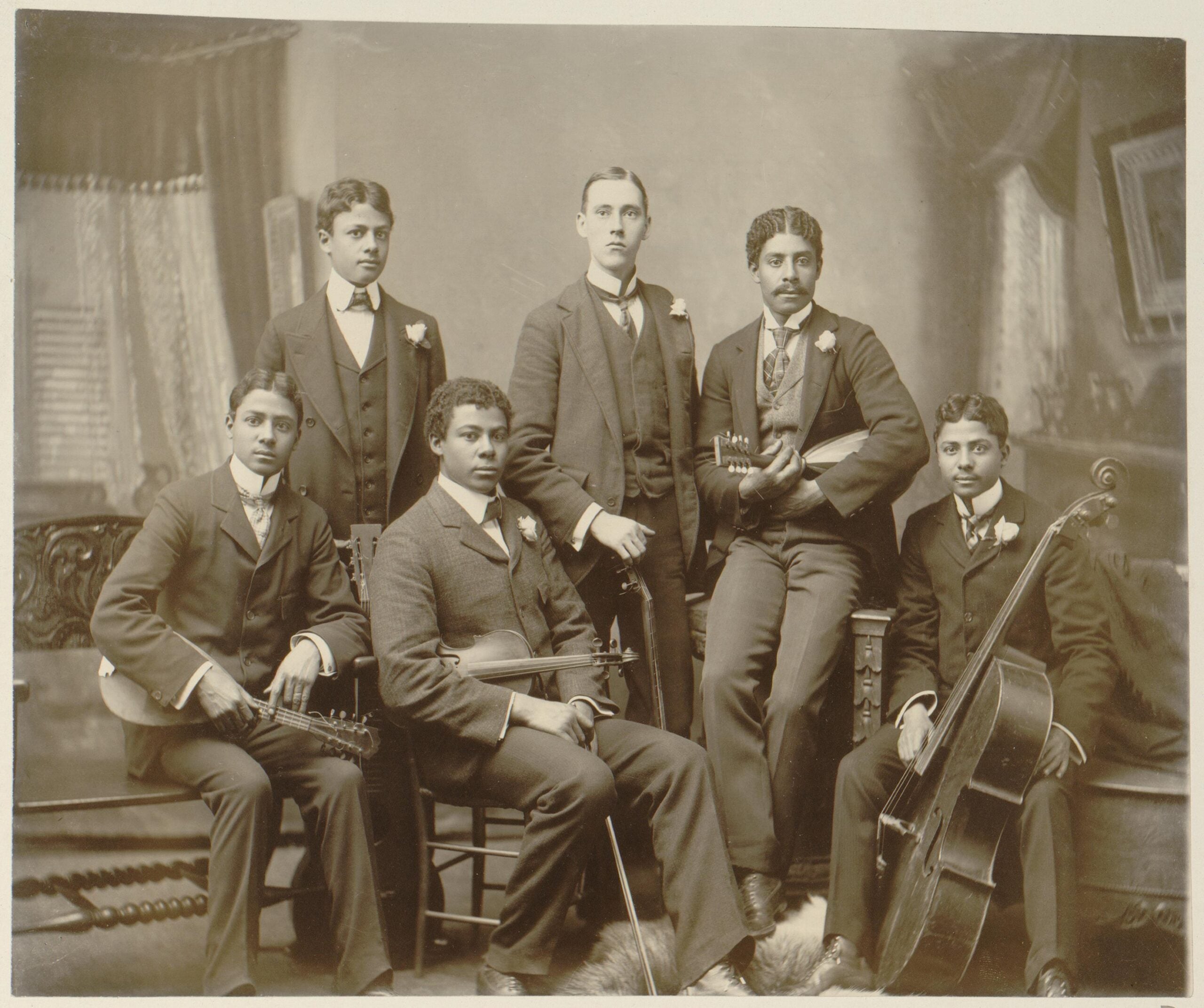 Six men in suits pose indoors with string instruments, including a cello, violin, and banjo. They sit and stand in a formal, vintage setting with draped curtains and framed pictures in the background.