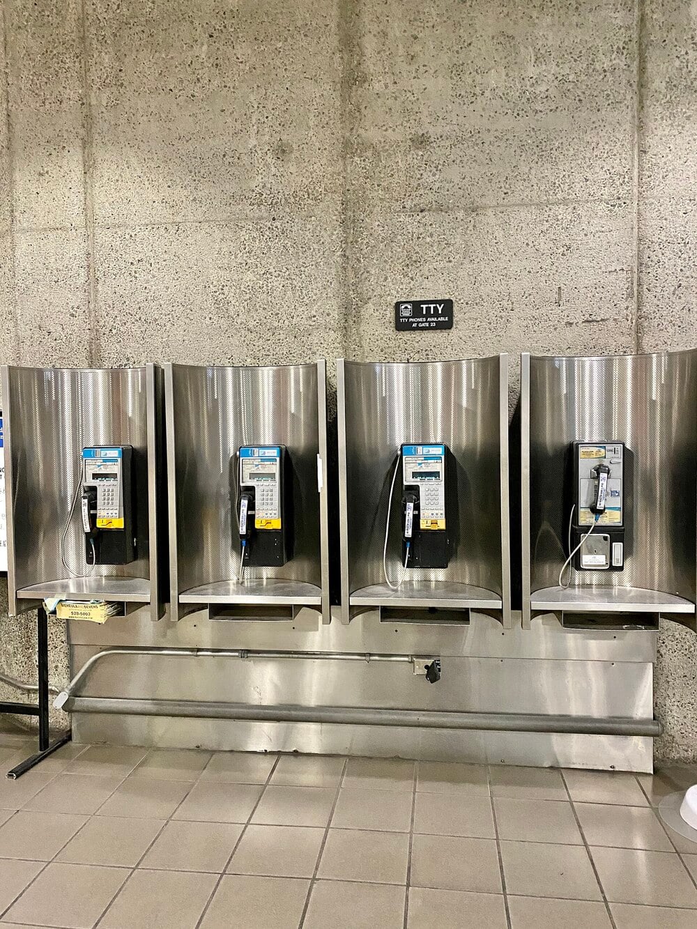 Four payphones with metal enclosures are mounted on a concrete wall. A sign above indicates TTY access. The floor is tiled, and exposed pipes run beneath the phones.