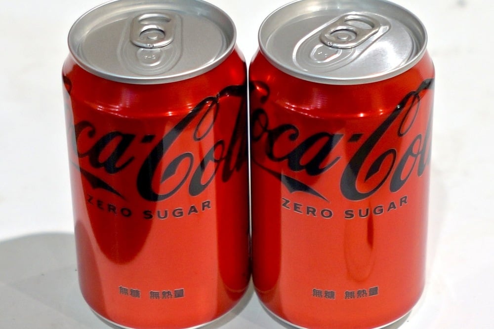 Two red cans of Coca-Cola Zero Sugar are placed side by side on a white surface. The cans feature the Coca-Cola logo in black with “Zero Sugar” written below it in English and additional text in another language.
