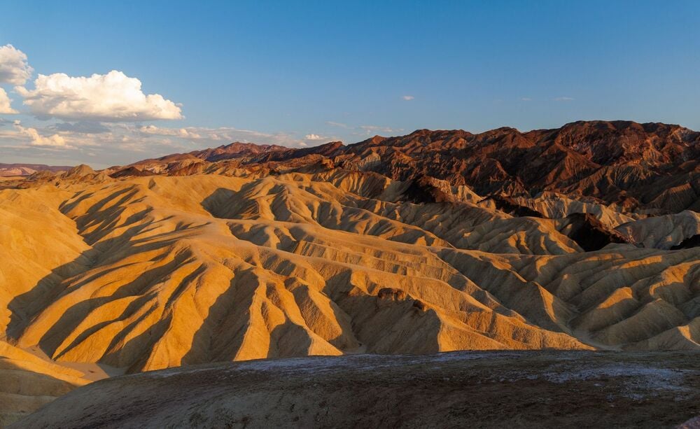 Golden, sunlit badlands with rippling ridges and eroded valleys stretch toward distant dark mountains under a clear blue sky with a few clouds.